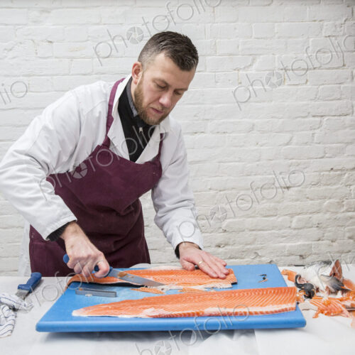 Man wearing apron standing at a table, cutting and filleting fresh salmon on blue… Man wearing apron standing at a table, cutting and filleting fresh salmon on blue…