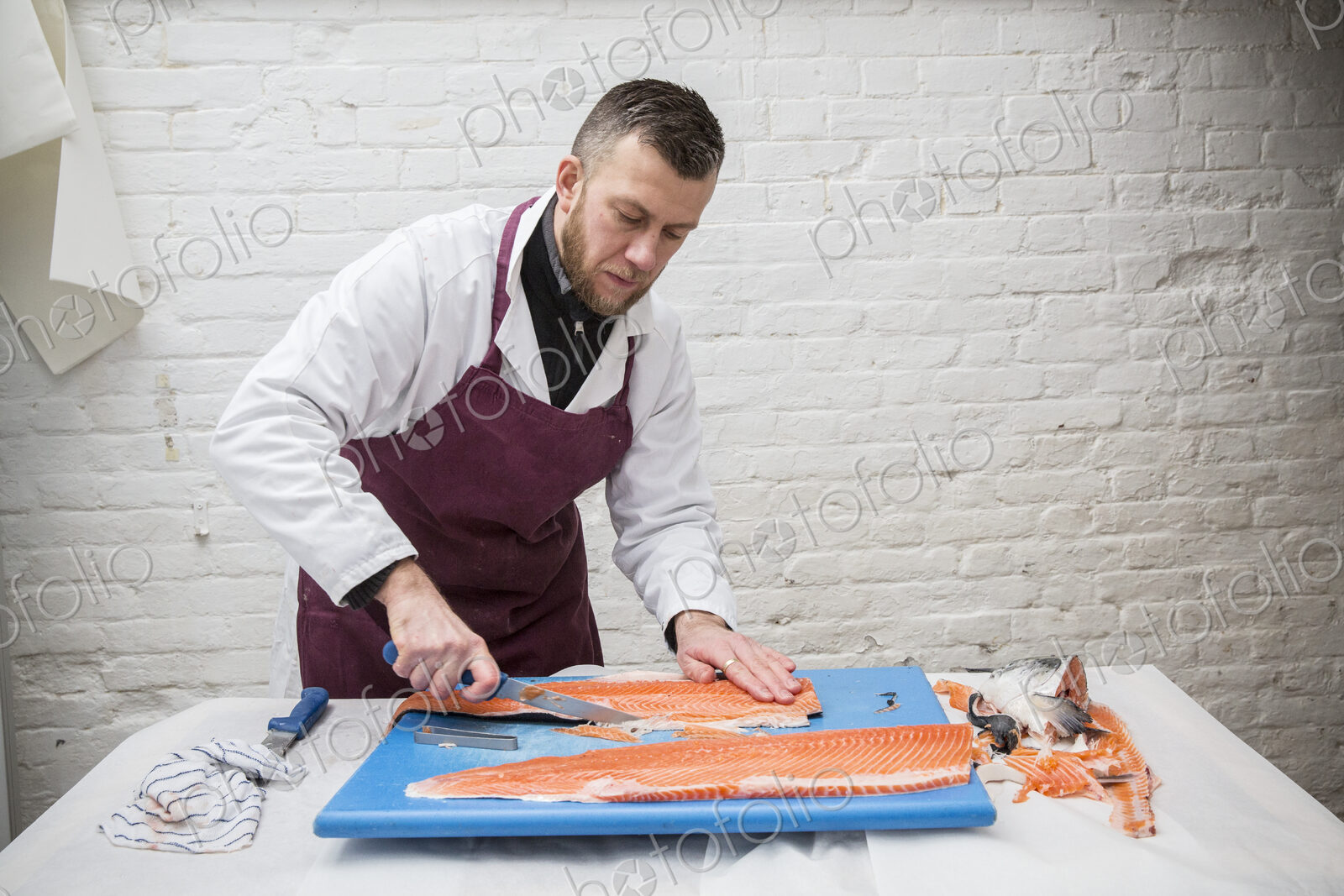 Man wearing apron standing at a table, cutting and filleting fresh salmon on blue…