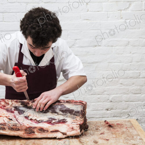 Man wearing apron standing at a wooden butcher's block, butchering beef forerib.