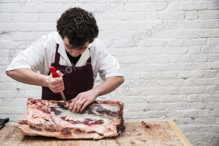 Man wearing apron standing at a wooden butcher's block, butchering beef forerib.