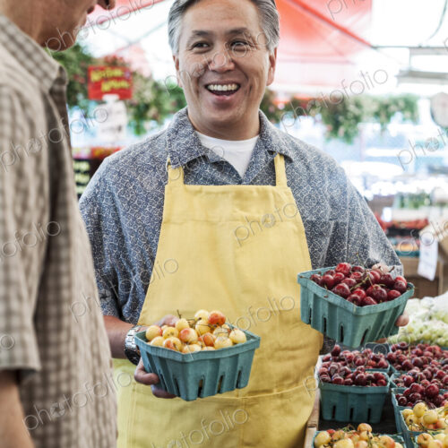 Man wearing apron standing at stall with punnets of fresh cherries at a fruit and… Man wearing apron standing at stall with punnets of fresh cherries at a fruit and…