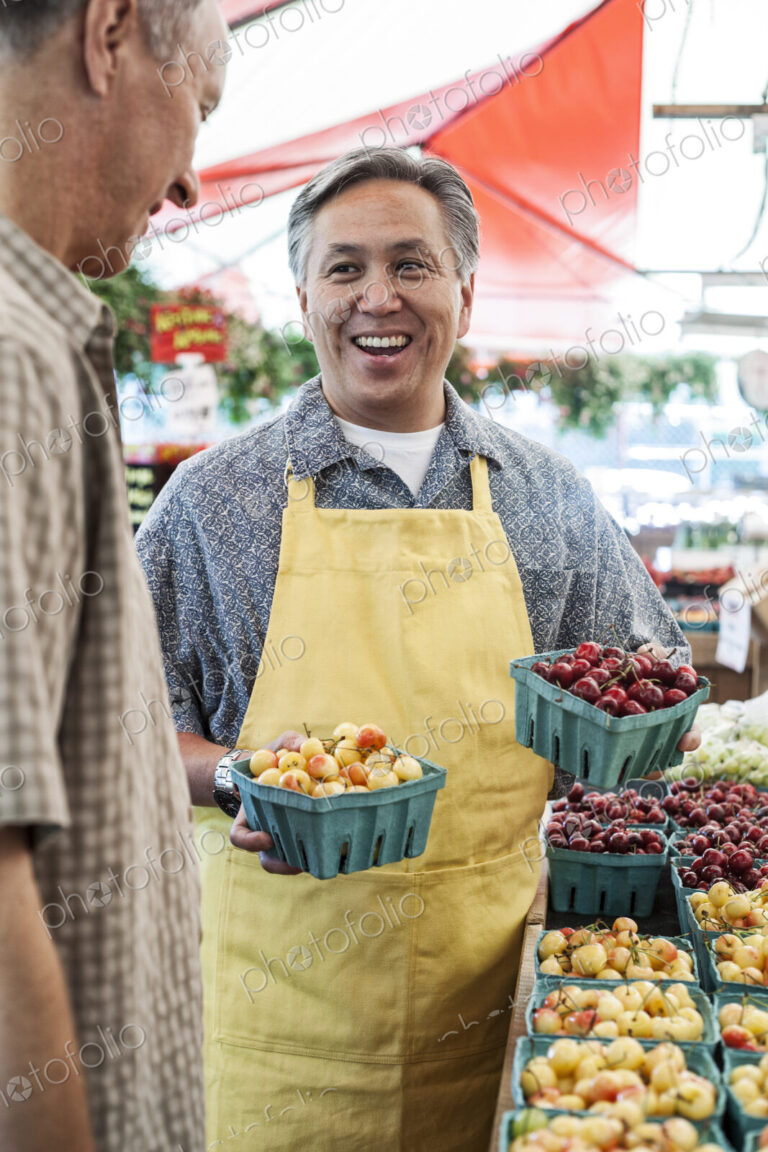 Man wearing apron standing at stall with punnets of fresh cherries at a fruit and…