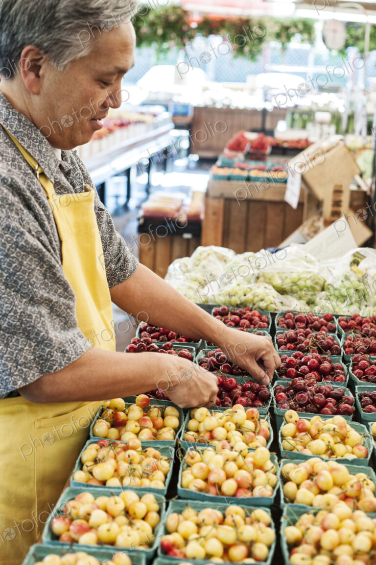 Man wearing apron standing at stall with punnets of fresh cherries at a fruit and…