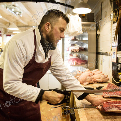Man wearing apron standing at wooden butcher's block, butchering piece of meat using saw.