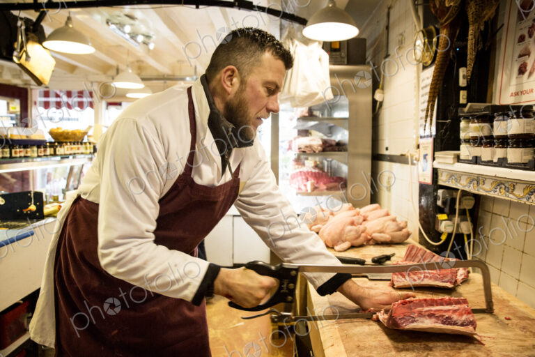 Man wearing apron standing at wooden butcher's block, butchering piece of meat using saw.