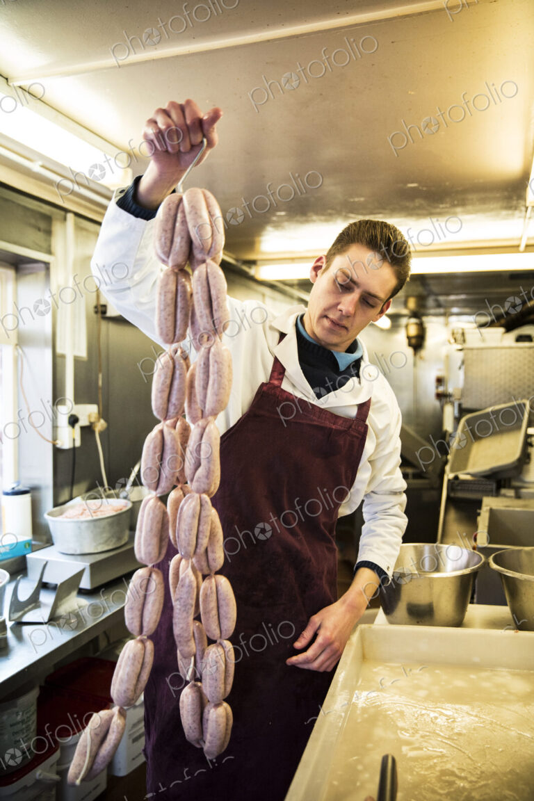 Man wearing apron standing in butcher shop, holding large number of freshly made…