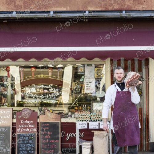 Man wearing apron standing on pavement outside butcher shop, carrying large piece of… Man wearing apron standing on pavement outside butcher shop, carrying large piece of…