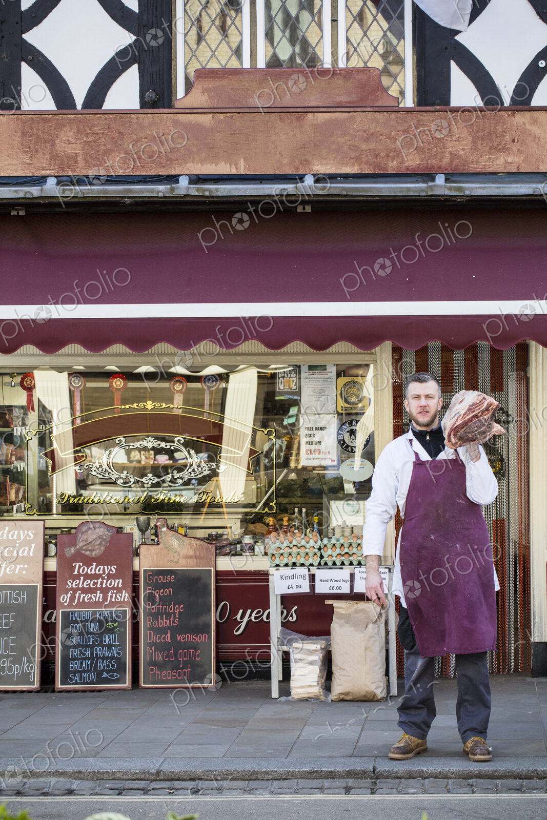 Man wearing apron standing on pavement outside butcher shop, carrying large piece of…