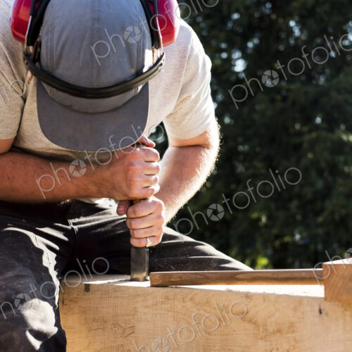 Man wearing baseball cap and ear protectors on building site, working on wooden beam.