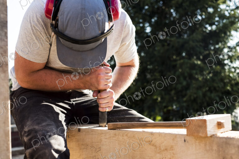 Man wearing baseball cap and ear protectors on building site, working on wooden beam.