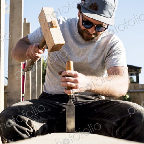 Man wearing baseball cap and sunglasses on building site, using mallet and chisel,…
