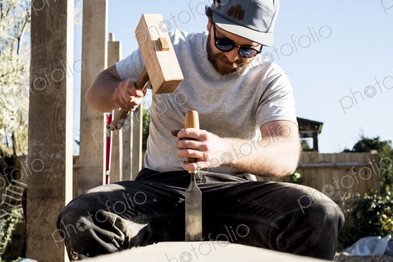 Man wearing baseball cap and sunglasses on building site, using mallet and chisel,…