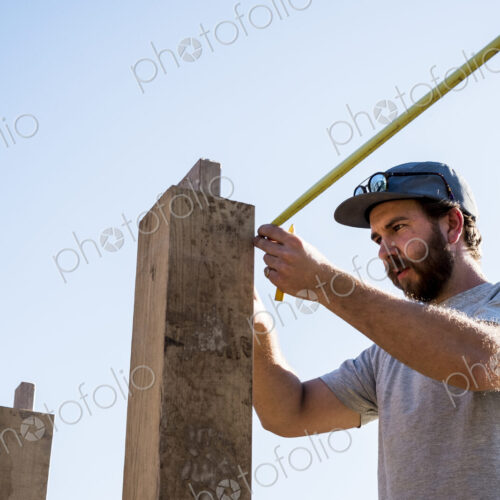 Man wearing baseball cap and sunglasses on building site, using tape measure.