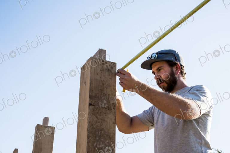 Man wearing baseball cap and sunglasses on building site, using tape measure.