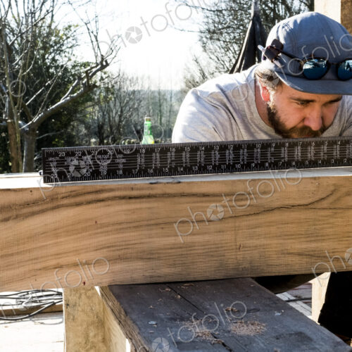 Man wearing baseball cap on building site, measuring wooden beam with metal ruler.