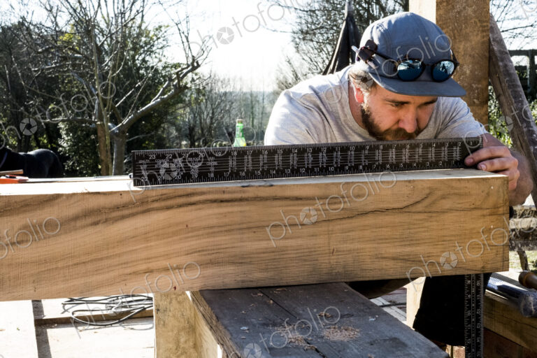 Man wearing baseball cap on building site, measuring wooden beam with metal ruler.