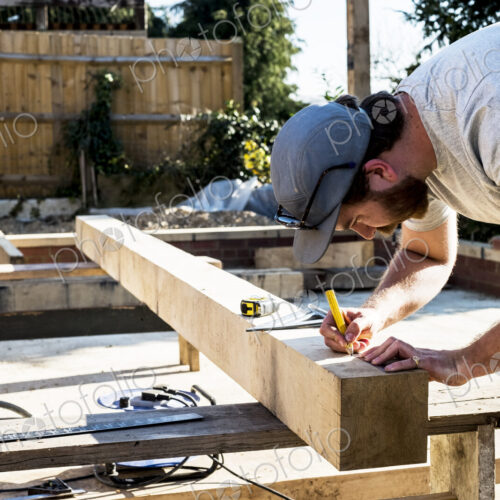 Man wearing baseball cap standing on building site, measuring and marking wooden beam.