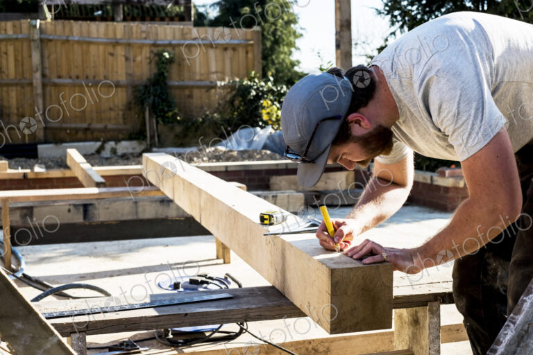 Man wearing baseball cap standing on building site, measuring and marking wooden beam.