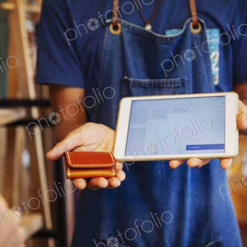 Man wearing blue apron and glasses standing in a leather shop, holding leather purse…