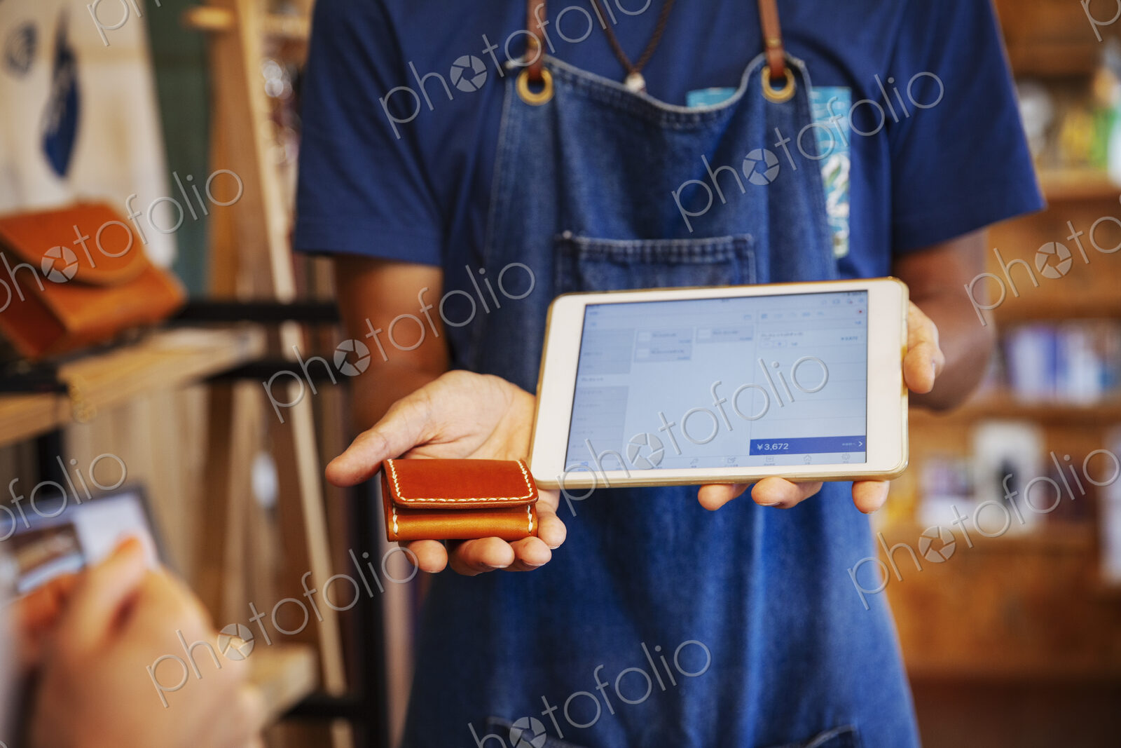 Man wearing blue apron and glasses standing in a leather shop, holding leather purse…
