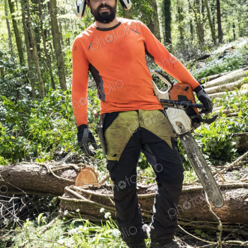 Man wearing bright orange top clearing part of forest. Cutting tree trunk with chain saw.