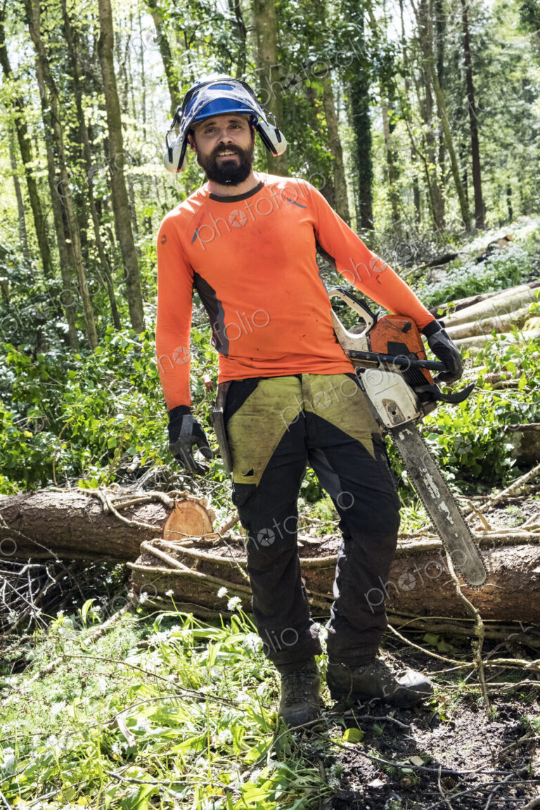 Man wearing bright orange top clearing part of forest. Cutting tree trunk with chain saw.