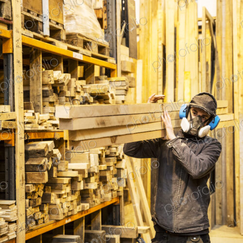 Man wearing ear protectors and dust mask standing in a warehouse, carrying long planks… Man wearing ear protectors and dust mask standing in a warehouse, carrying long planks…