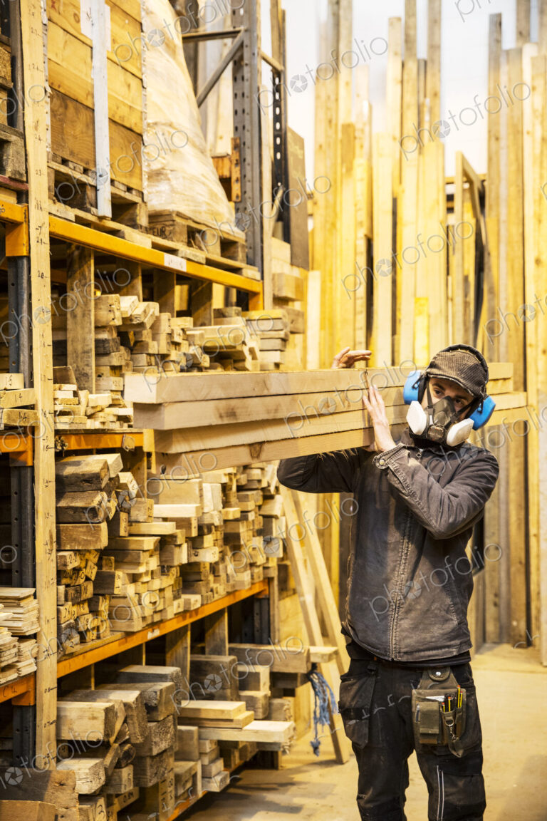 Man wearing ear protectors and dust mask standing in a warehouse, carrying long planks…