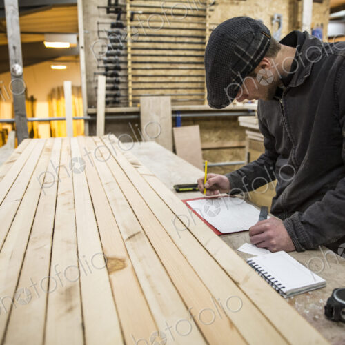 Man wearing flat cap standing at a workbench with wooden planks in a warehouse,… Man wearing flat cap standing at a workbench with wooden planks in a warehouse,…