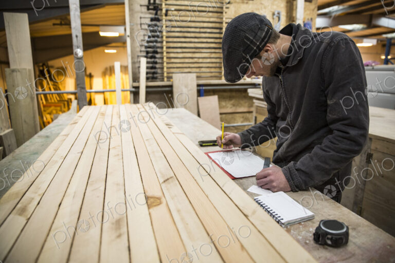 Man wearing flat cap standing at a workbench with wooden planks in a warehouse,…