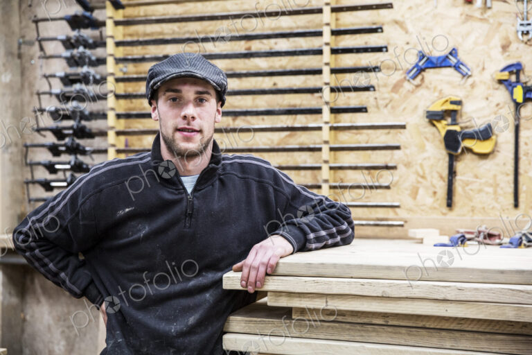Man wearing flat cap standing next to a stack of wooden boards, looking at camera,…