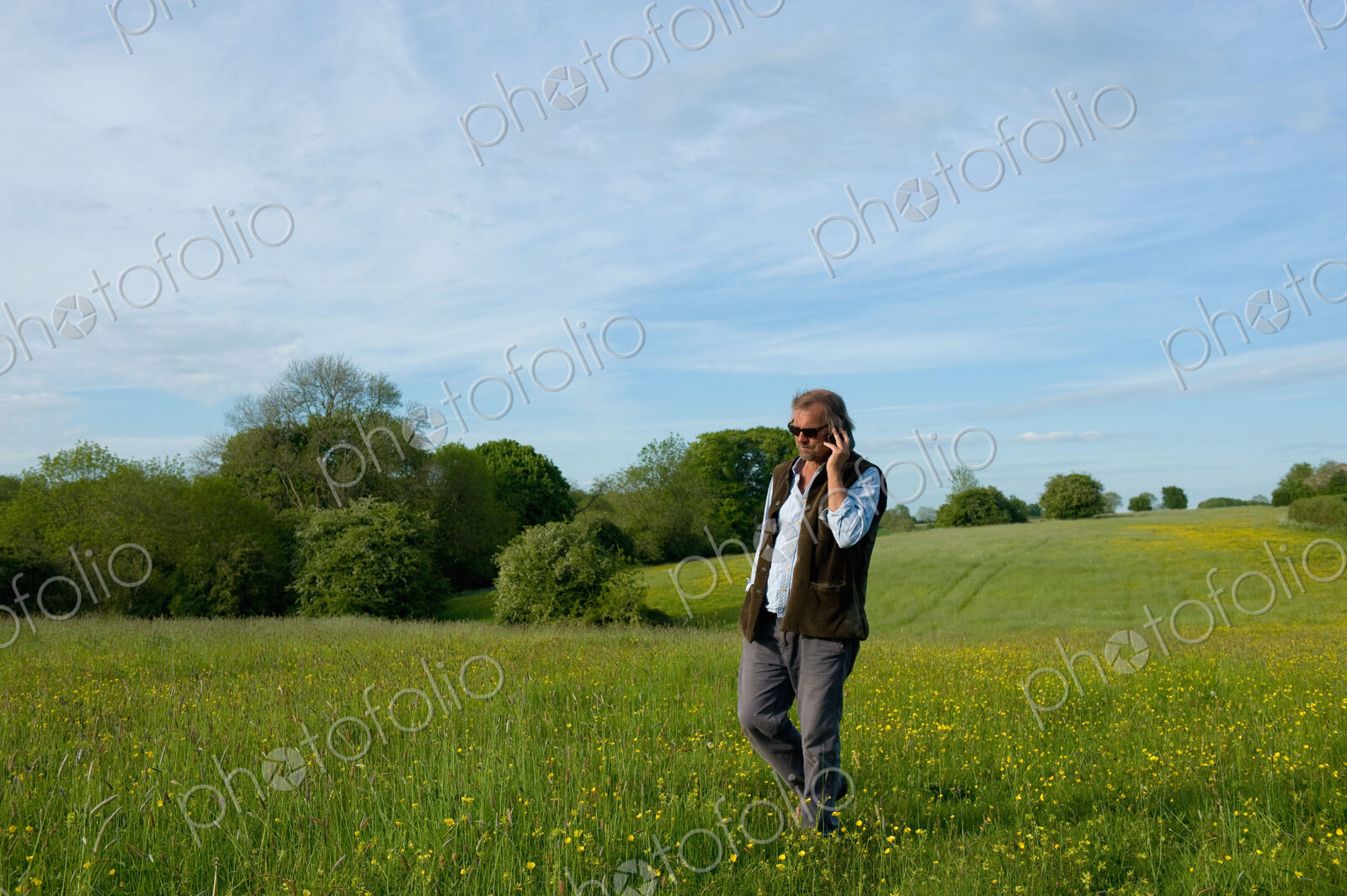 Man wearing sunglasses walking across meadow, holding mobile phone to his ear.