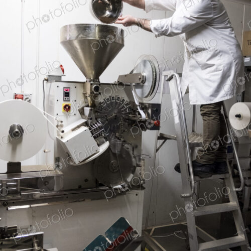 Man wearing white coat standing on a ladder, pouring tea leaves into large funnel of a…