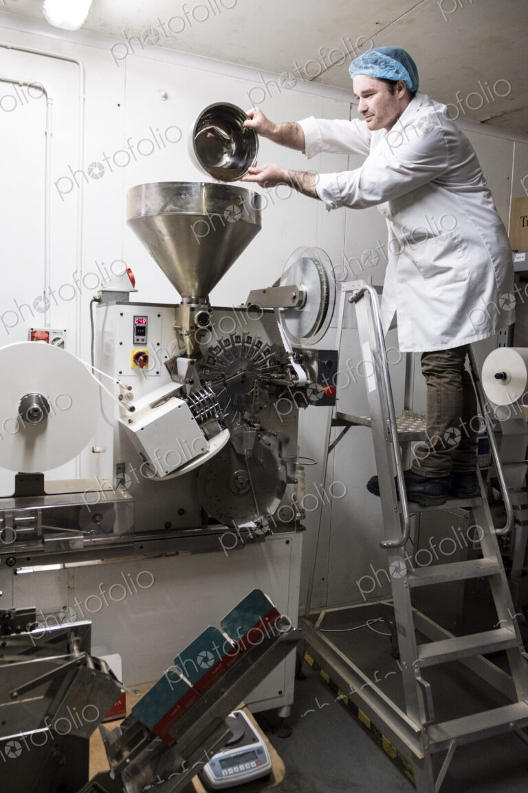Man wearing white coat standing on a ladder, pouring tea leaves into large funnel of a…