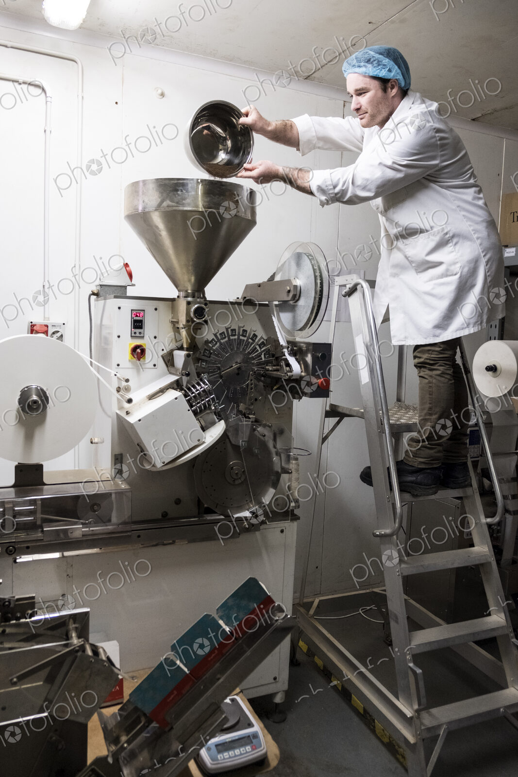 Man wearing white coat standing on a ladder, pouring tea leaves into large funnel of a…