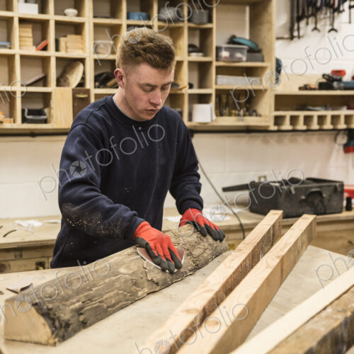Man wearing work gloves standing at a workbench in a workshop, sanding bark of a piece… Man wearing work gloves standing at a workbench in a workshop, sanding bark of a piece…