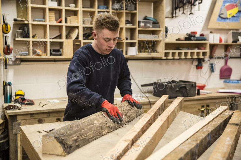 Man wearing work gloves standing at a workbench in a workshop, sanding bark of a piece…