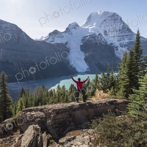 Man with arms raised, on a ridge above Berg lake and mountains.