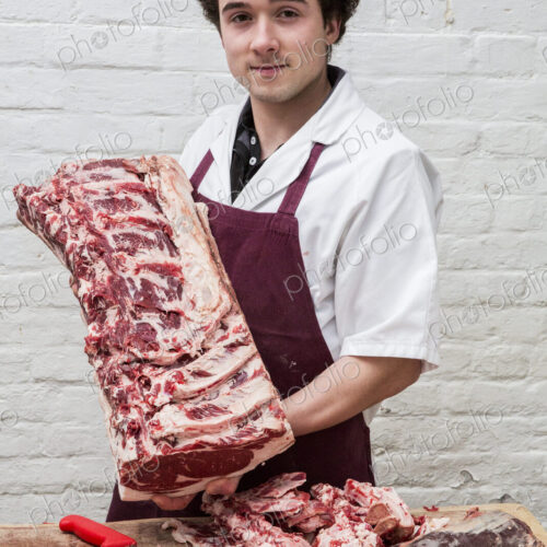 Man with brown curly hair wearing apron standing at a wooden butcher's block, holding…