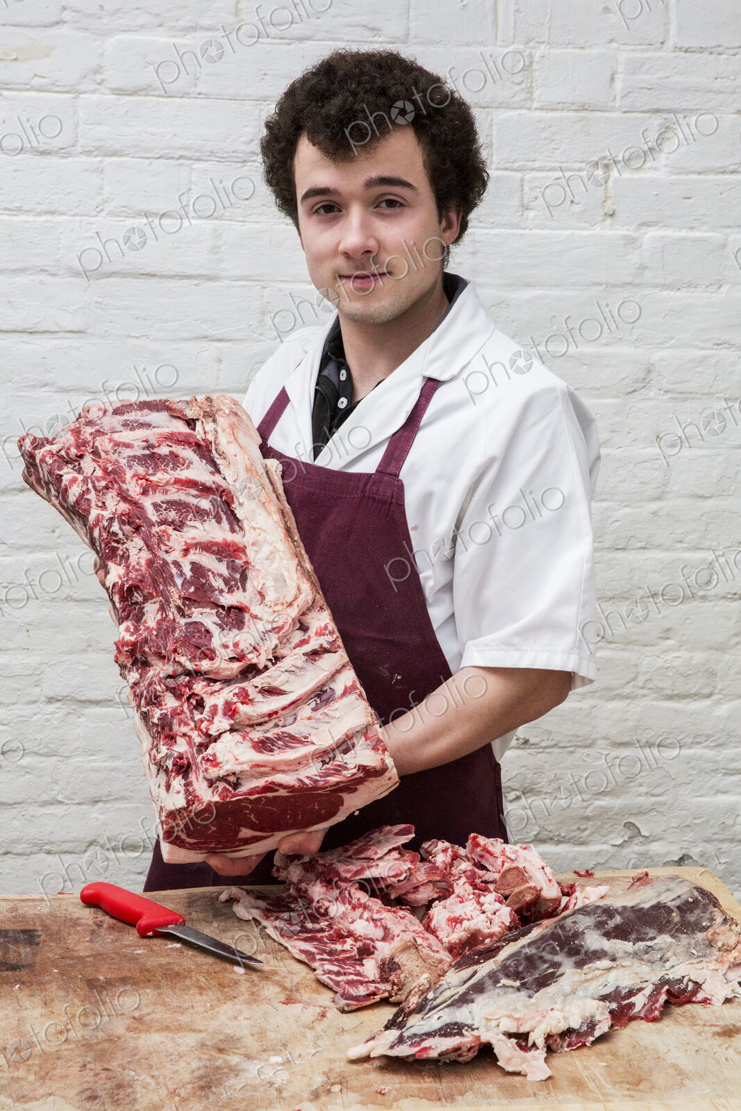Man with brown curly hair wearing apron standing at a wooden butcher’s block, holding…