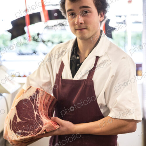 Man with brown curly hair wearing apron standing in butcher shop, holding beef… Man with brown curly hair wearing apron standing in butcher shop, holding beef…