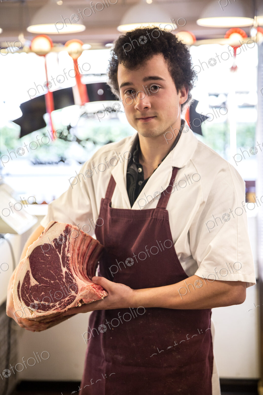 Man with brown curly hair wearing apron standing in butcher shop, holding beef…