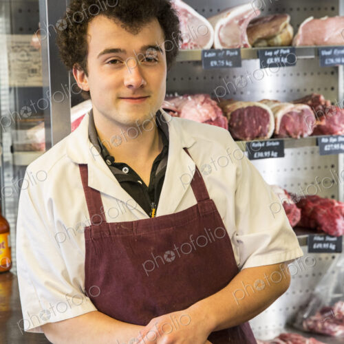 Man with brown curly hair wearing apron standing in butcher shop, smiling at camera.
