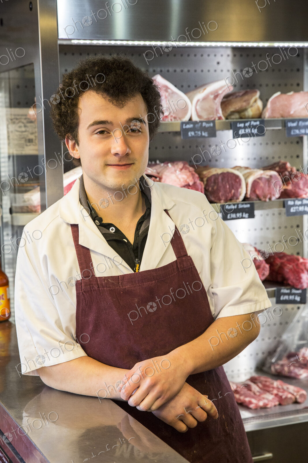 Man with brown curly hair wearing apron standing in butcher shop, smiling at camera.