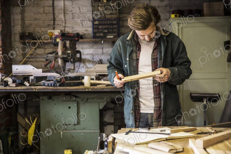 Man working standing at a workbench in a woodworking workshop, holding piece of wood.
