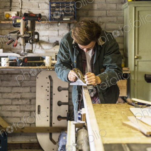 Man working standing at a workbench in a woodworking workshop, using plane on plank of…