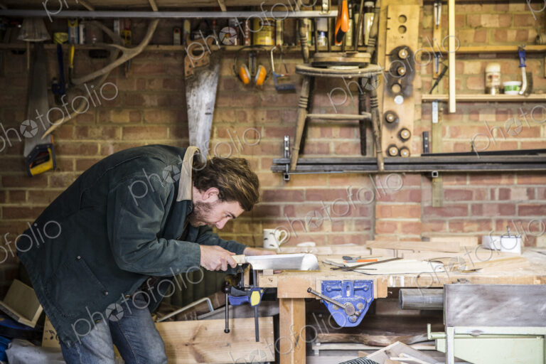 Man working standing at a workbench in a woodworking workshop, using saw on piece of wood.