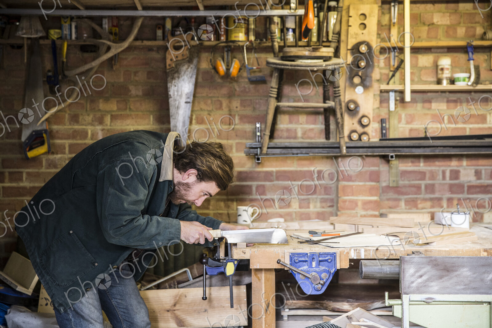 Man working standing at a workbench in a woodworking workshop, using saw on piece of wood.