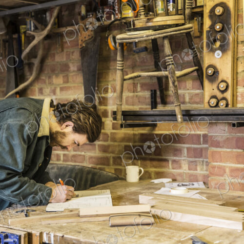 Man working standing at a workbench in a woodworking workshop, writing in notebook.
