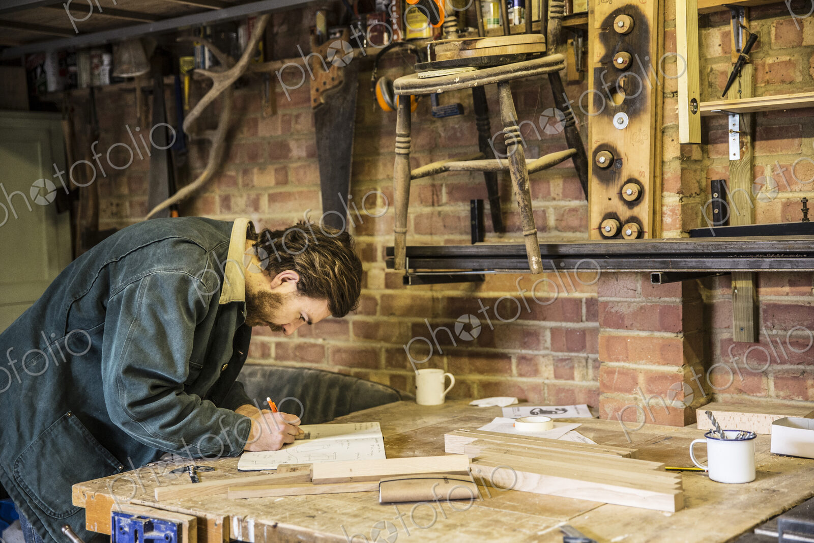 Man working standing at a workbench in a woodworking workshop, writing in notebook.